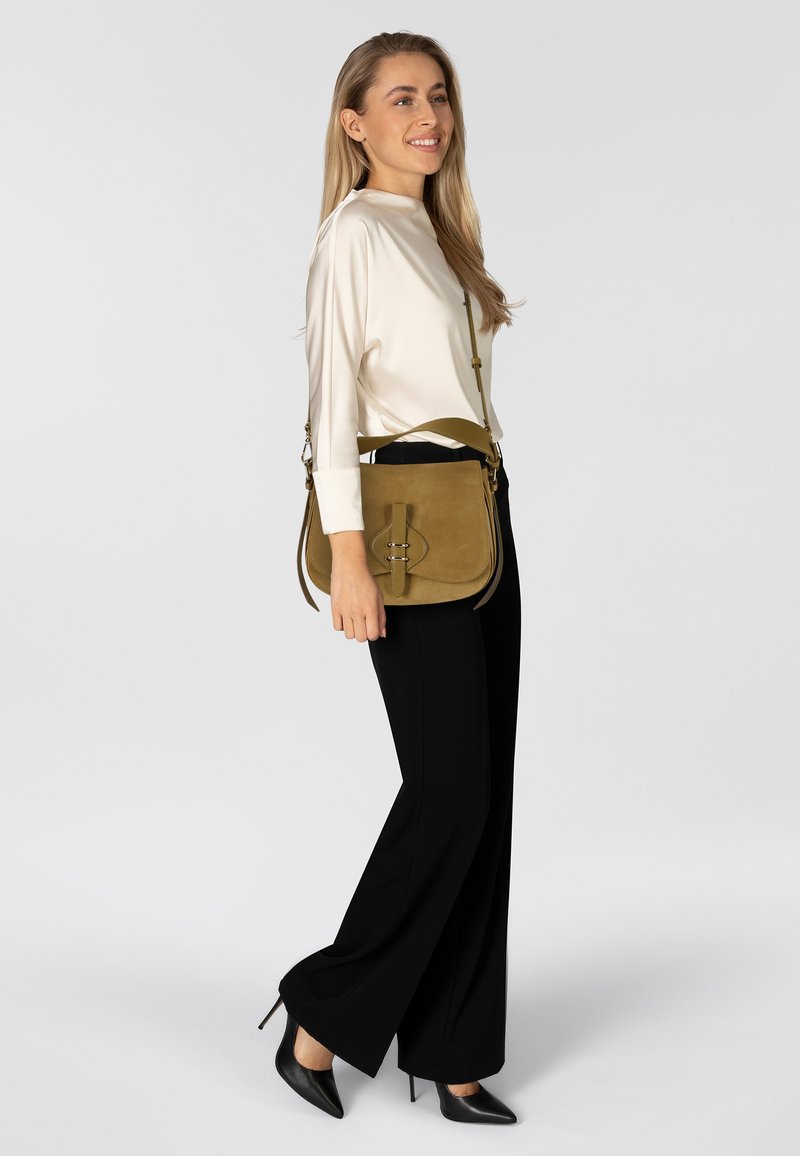 Borsa a tracolla in camoscio verde oliva con dettaglio di chiusura frontale, abbinata a una camicetta color crema e pantaloni neri a gamba larga.