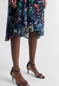 Blommönstrad, genomskinlig mörk klänning med starka färger; kombinerad med svarta högklackade sandaler med remmar och silverdetaljer.