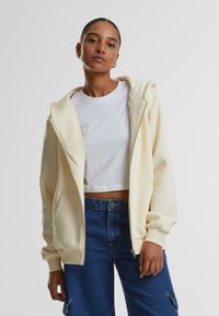 Crème kleurige zip-up hoodie met een zachte textuur en een capuchon, gedragen over een wit cropped T-shirt en gecombineerd met hoog-waist blauwe denim broeken.