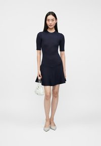 Vestido de punto en navy de manga corta con un cuerpo ajustado y falda acampanada. La modelo lleva un pequeño bolso blanco y usa zapatos de tacón en gris claro.
