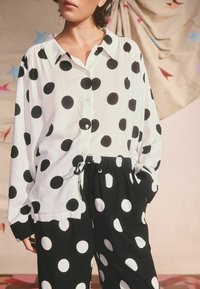 Čierna a biela košeľa s bodkami, s golierom a gombíkmi, v kombinácii s rovnakými čiernymi nohavicami. Mäkko tkaný materiál, voľný strih a pás na šnurovanie.