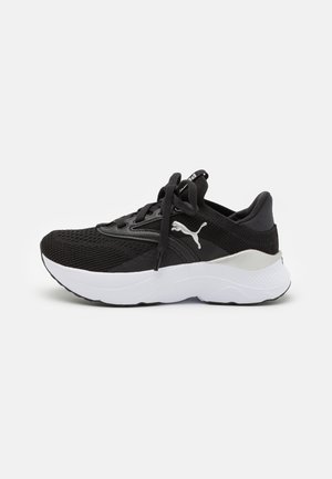 Zapatilla deportiva negra con parte superior de malla, entresuela blanca, puntera redonda y diseño de cordones. Presenta un logo prominente en el lateral y acentos texturizados.