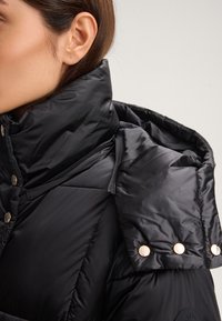 Giubbotto nero imbottito con colletto alto, realizzato in tessuto lucido, design delle spalle arrotondati e bottoni a pressione dorati come dettagli distintivi.