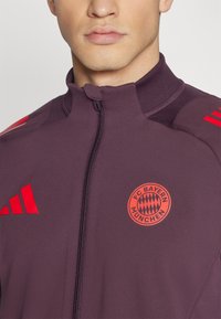 FC Bayern Miunchen striukė tamsiai purpurinės spalvos audinyje. Yra raudonos akcentai ir logotipas, aukšta apykaklė ir pilnas užtrauktukas. Sklandus paviršius su sportiniu dizainu.