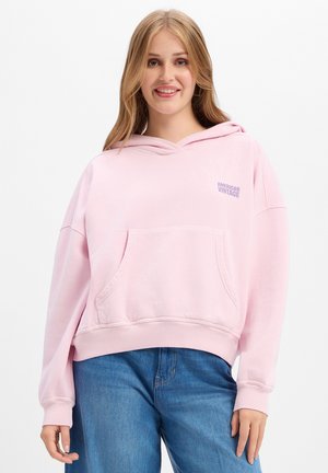 Jonge vrouw met lang blond haar, gedragen een lichtroze hoodie met de tekst "American Vintage" en een blauwe spijkerbroek, glimlachend en staand tegen een witte achtergrond.