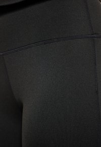 Fekete, rugalmas leggings sima textúrával és lapos varrásokkal. Az anyag nedvességel lélegzőnek tűnik és testhez simul, alakot formálva.