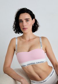 Ružičasti bralet s glatkom teksturom, s prilagodljivim bijelim trakama i bijelom trakom s crnim logotipom "Calvin Klein" duž dna.