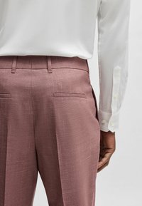 Aangepaste broek van licht mauve stof met een gestructureerde afwerking. Bevat twee achterzakken en een plooi aan de voorkant. Gecombineerd met een wit shirt.