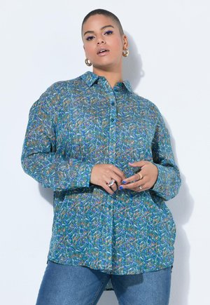 Frau mit kurzen Haaren, die ein blau gemustertes Hemd mit Knöpfen, goldene Creolen und Jeans trägt und vor einem weißen Hintergrund posiert.