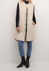 Beżowa pikowana kamizelka z wysokim kołnierzem, z zatrzaskami, dwoma bocznymi kieszeniami i luźnym krojem, noszona na granatowej bluzie z długim rękawem i niebieskich dżinsach.