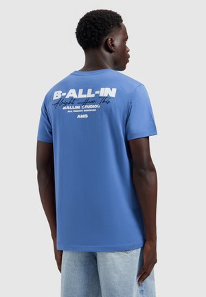 Man draagt een blauw T-shirt met witte tekst "B-ALL-IN" en extra kleinere tekst op de achterkant, gecombineerd met lichtblauwe jeans.