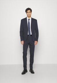 Marineblauer Anzug mit zwei Knöpfen, mit Revers und seitlichen Taschen. Kombiniert mit einem weißen Hemd und einer dunklen Krawatte. Die Schuhe sind aus schwarzem Leder.