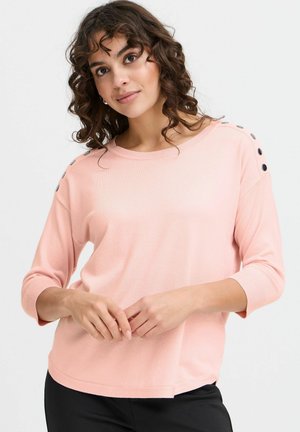 Haut rose clair à manches longues en tissu doux. Présente un col arrondi et des boutons noirs décoratifs sur les épaules. Coupe décontractée.