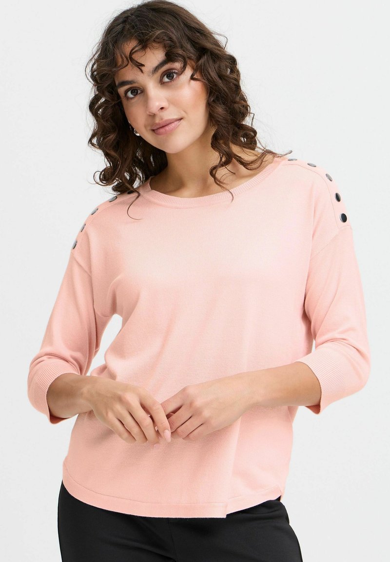 Haut rose clair à manches longues en tissu doux. Présente un col arrondi et des boutons noirs décoratifs sur les épaules. Coupe décontractée.