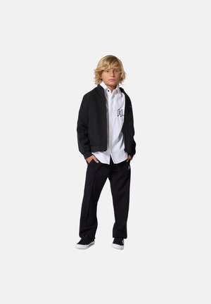 Zwarte bomberjack over een wit shirt met geborduurd logo, gecombineerd met zwarte broek en zwarte sneakers. Kort, golvend blond haar.