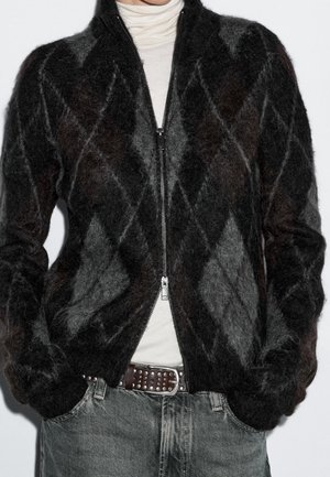 Personne portant un pull à fermeture éclair argyle noir et gris duveteux sur un col roulé blanc, un jean gris et une ceinture marron cloutée.