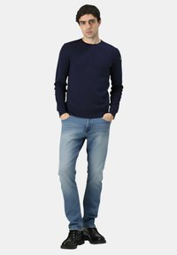 Maglione in maglia blu navy con scollo rotondo, abbinato a jeans di denim azzurro chiaro. La modella indossa stivaletti neri alla caviglia, con le mani in tasca.