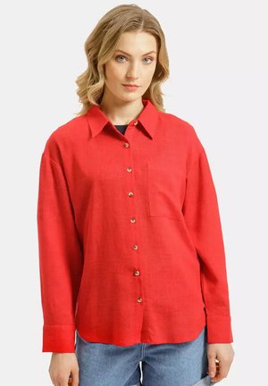 Kobieta ubrana w jasnoczerwoną długą koszulę z guzikiem, z kieszenią z przodu, oraz jasnoniebieskie dżinsy, stojąca na jednolitym tle.