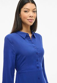Modrá blúzka s dlhými rukávmi, golierom a hladkou textúrou. Obsahuje radu zladených gombíkov na prednej strane.