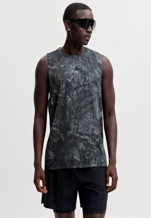 Homme portant un débardeur de sport noir et gris, un short noir, de grandes lunettes de soleil noires et des boucles d'oreilles en or, debout devant un fond uni.