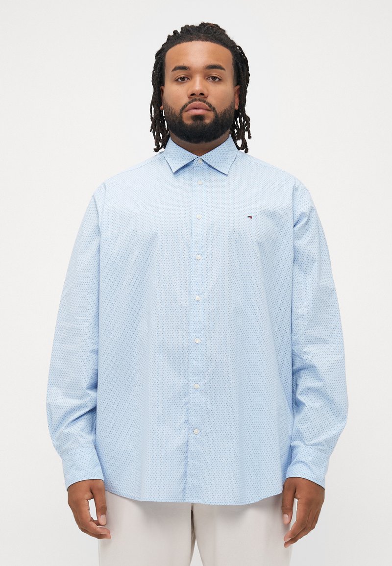 Hombre con rastas vistiendo una camisa de botones azul claro con estampado y pantalones beige, de pie contra un fondo blanco liso.