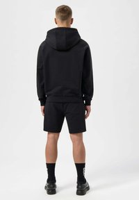Black hooded sweatshirt with a relaxed fit, paired with black shorts. Material appears soft, with ribbed cuffs and waistband. Black socks and shoes.