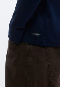 Μακρυμάνικο πουκάμισο σε ναυτικό μπλε με υφή υφάσματος, που φέρει μια ετικέτα με λογότυπο στο κάτω μέρος, σε συνδυασμό με καφέ παντελόνι από κοτλέ.