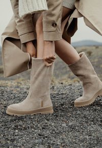 Botas de ante beige hasta la rodilla con un interior suave, que cuentan con una suela texturizada y una pequeña etiqueta de logo. Colocadas sobre un fondo de grava.