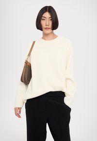 Pull crème à côtes avec un col rond, assorti à un pantalon noir. Un sac à main à motif marron est porté à l'épaule.