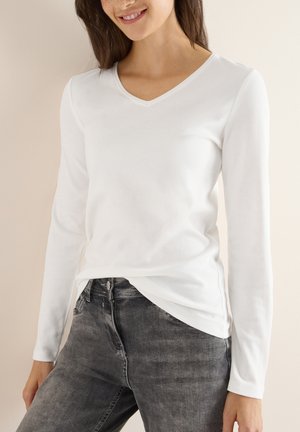 T-shirt à col V blanc à manches longues en tissu doux, avec un ourlet droit et une silhouette légèrement ajustée, associé à un jean gris.
