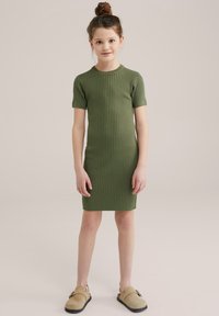 Žalia juostuota mezgimo suknelė su trumpomis rankovėmis, prigludęs dizainas, iki kelių siekianti apdaila, derinama su smėline slip-on tipo avalyne ir paprasta šukuosena.