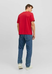 Rode katoenen T-shirt met een klassieke ronde hals; gecombineerd met wijde blauwe spijkerbroek en witte sneakers, gezien vanuit de achterkant.