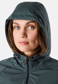 Veste à capuche en tissu vert sarcelle foncé, avec une fermeture éclair à l'avant, un capuchon à cordon de serrage et des détails de couture visibles. Texture lisse avec un design ajusté.