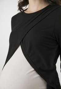 Top gravidanza nero con design incrociato, tessuto elastico, maniche lunghe e orlo curvo, sovrapposto a uno strato sottostante di colore chiaro.