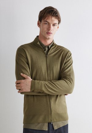 Jeune homme à la peau claire et aux cheveux bruns debout, les bras croisés, portant un pull zippé vert olive par-dessus une chemise devant un mur uni.