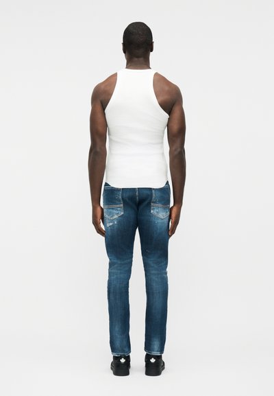 Άντρας που στέκεται γυρισμένος με την πλάτη, φορώντας άσπρο αμάνικο μπλουζάκι, μπλε τζιν με πίσω τσέπες και μαύρα αθλητικά παπούτσια, μπροστά σε ένα απλό φόντο.