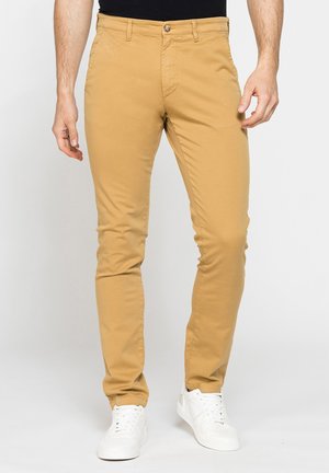 Uomo con pantaloni slim color senape e sneaker bianche, in piedi davanti a uno sfondo chiaro e uniforme.