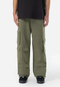 Pantaloni cargo din bumbac de culoare verde măslin, cu numeroase buzunare mari, croi lejer și cusături întărite. Purtați cu un tricou negru și pantofi negri.
