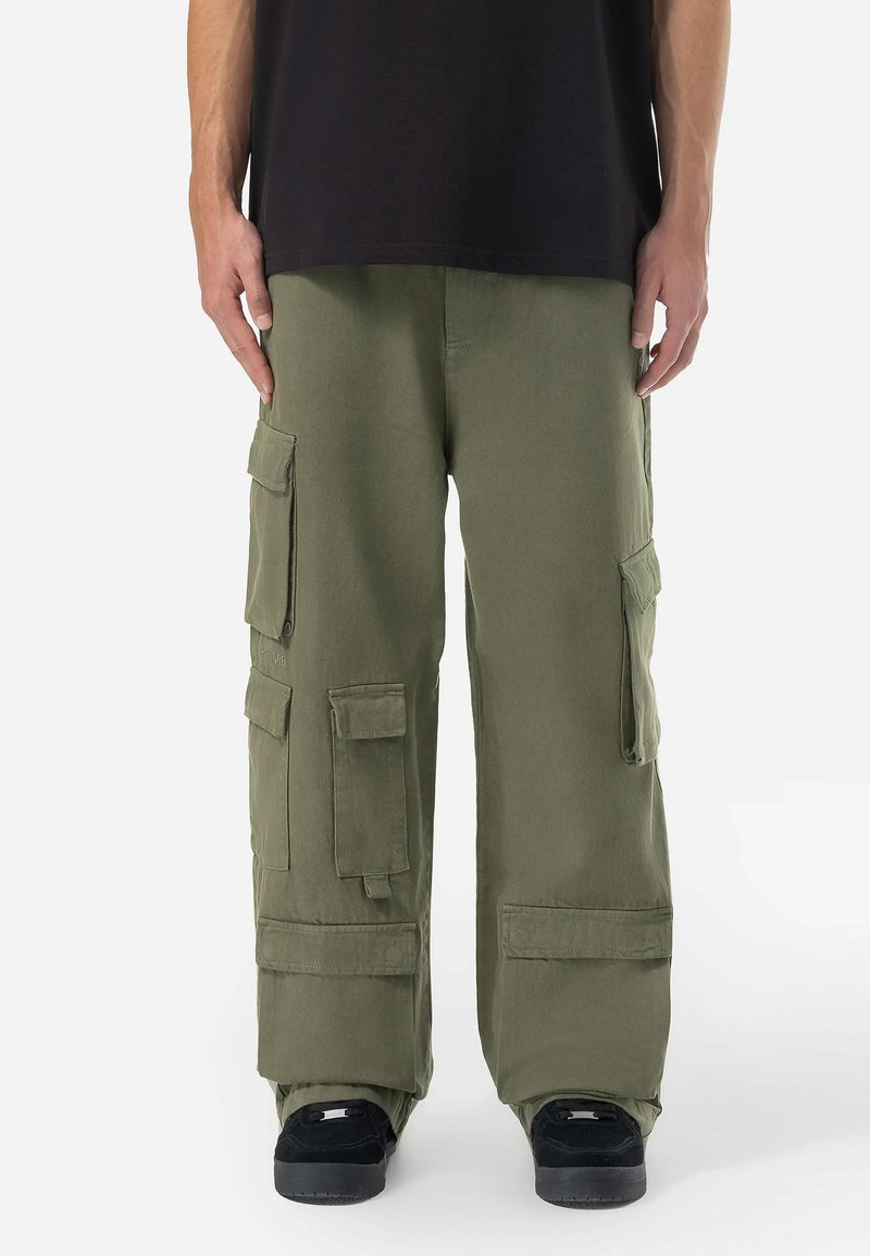 Pantaloni cargo din bumbac de culoare verde măslin, cu numeroase buzunare mari, croi lejer și cusături întărite. Purtați cu un tricou negru și pantofi negri.