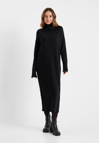 Zwarte gebreide jurk met hoge kraag, lange mouwen en een relaxed fit, met ribbelstructuur bij de mouwen en de zoom. Gecombineerd met zwarte laarzen.