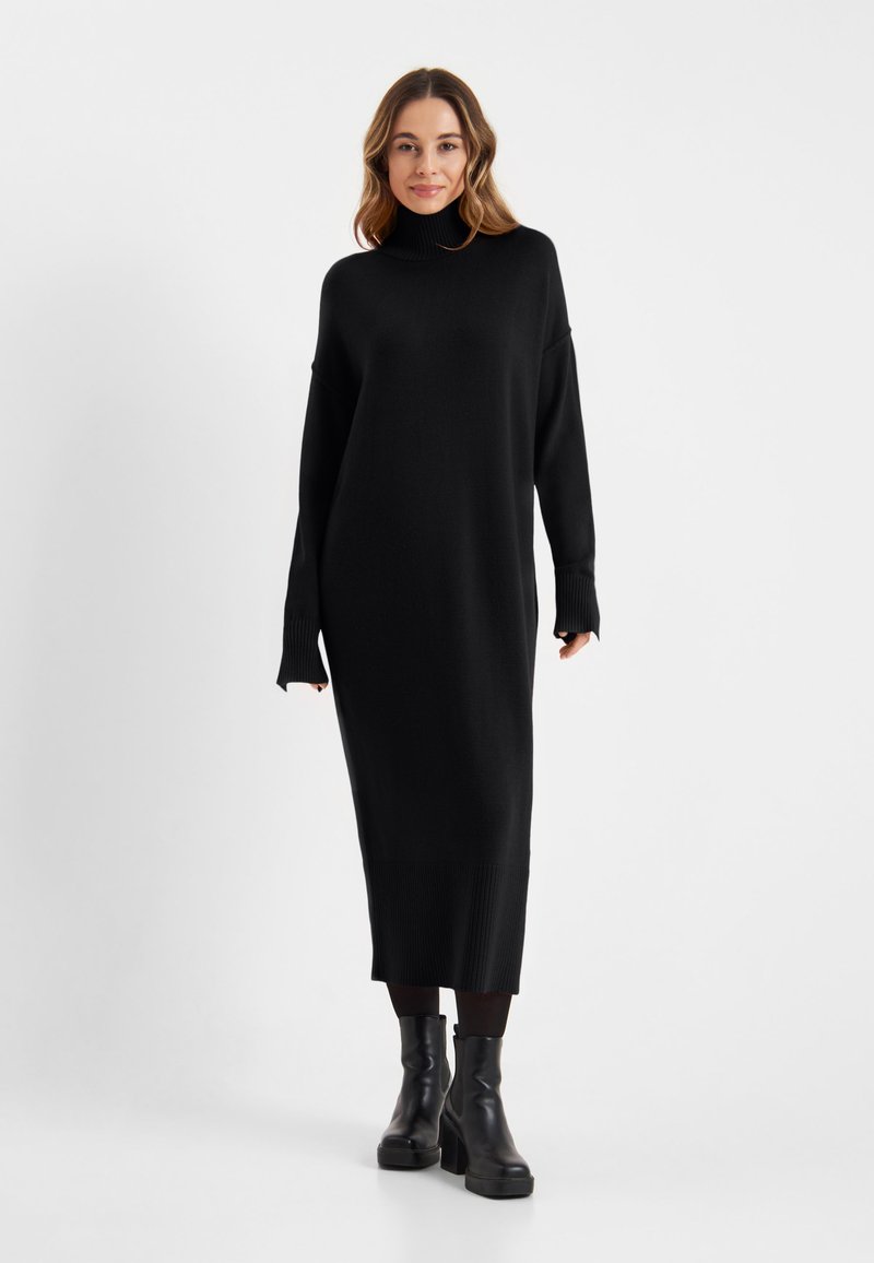 Zwarte gebreide jurk met hoge kraag, lange mouwen en een relaxed fit, met ribbelstructuur bij de mouwen en de zoom. Gecombineerd met zwarte laarzen.