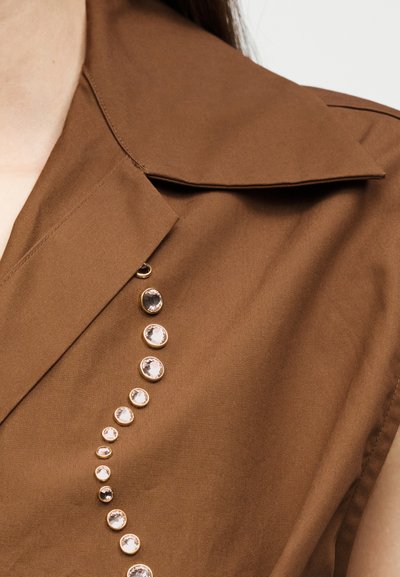 Gros plan sur un vêtement marron sans manches avec col pointu et rangée verticale de pierres précieuses rondes transparentes décoratives.