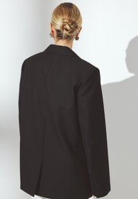 Blondide juustega naine madala kuklaga, kes on musta ülikonna seljas ja seisab seljaga, lihtsa heledat tausta ees, millele on seinale langenud vari.