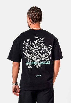 Mann mit geflochtenem Haar, der ein schwarzes T-Shirt mit weißem Blumendesign und dem Schriftzug "Garden Energy" auf der Rückseite trägt.