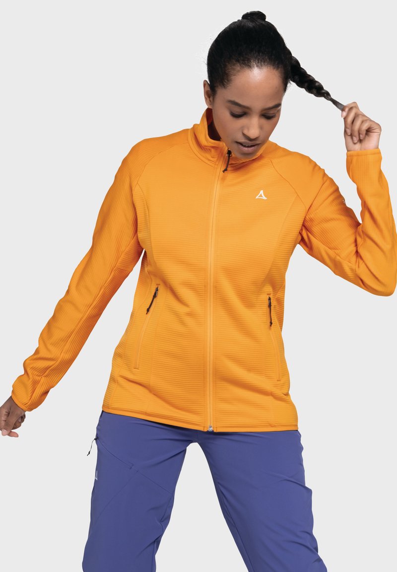 Orange Zip-Jacke mit geripptem Muster, ausgestattet mit zwei Seitentaschen und einem Logo auf der Brust. Getragen mit lila Hosen.