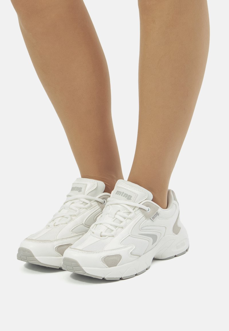 Zapatillas deportivas blancas con una parte superior texturizada, detalles en gris y una suela acolchada, que cuentan con cordones y un logotipo en la lengüeta.
