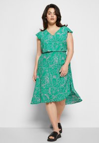 Vestido verde com mangas curtas em babados, decote em V, cintura ajustada, apresentando um padrão floral em rosa e branco. Usado com sandálias pretas.