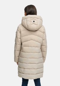 Vrouw in een beige gewatteerde knielange pufferjack met capuchon, staande met haar rug naar de camera tegen een effen achtergrond.