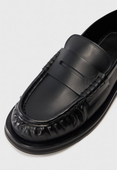 Melna ādas penny loafers ar savāktu apdari ap purngalu un gludu joslu pāri pēdas augšai uz plakana zole.