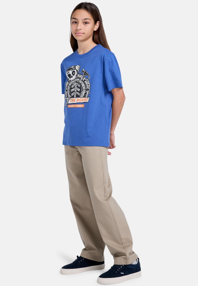 Blå bomulds T-shirt med grafikprint, der viser et kranium og værktøj, parret med beige bukser og mørkeblå sneakers.